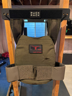 Olive green GORUCK plate carrier with a brand patch, hanging on a black Ruck Rack wall-mounted storage rack for tactical gear organization.