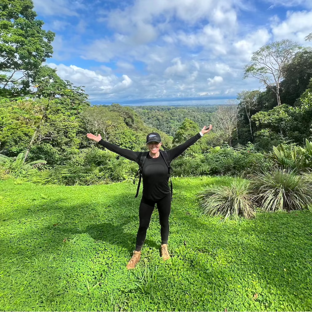 Person in black outfit stands on lush grass with arms wide, lush forest and blue sky in the background.