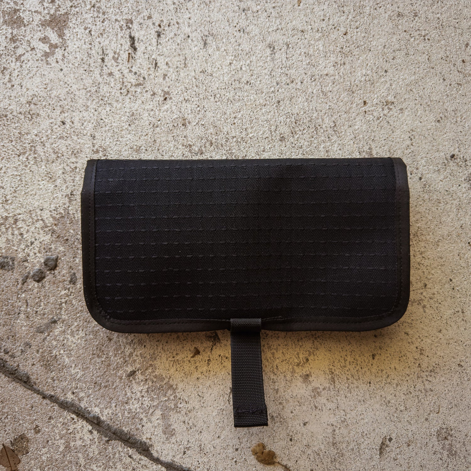A closed Scars Wire Dopp, an EDC pouch with a strap made from black waxed twill, rests on a textured concrete surface.