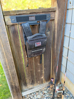 Black GORUCK rucksack hanging on a wall-mounted storage rack attached to a wooden gate, with fishing rods leaning against the side for tactical gear organization.