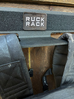 A black metal wall-mounted gear hanger with a “RUCK RACK” patch, used for tactical gear organization while holding the shoulder straps of a black backpack.