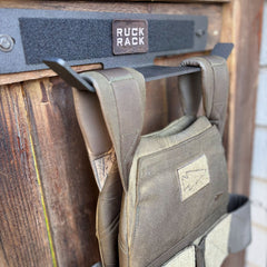An olive green tactical weight vest hangs on a black metal wall-mounted storage rack designed for tactical gear organization.