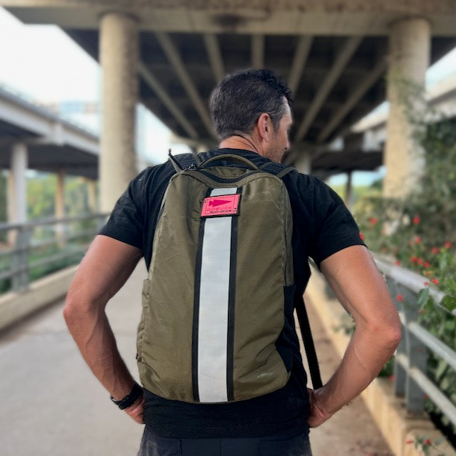 A person wearing an olive green rucksack with a vertical reflective stripe and a small pink tag, seen from behind while rucking on a path under an overpass.