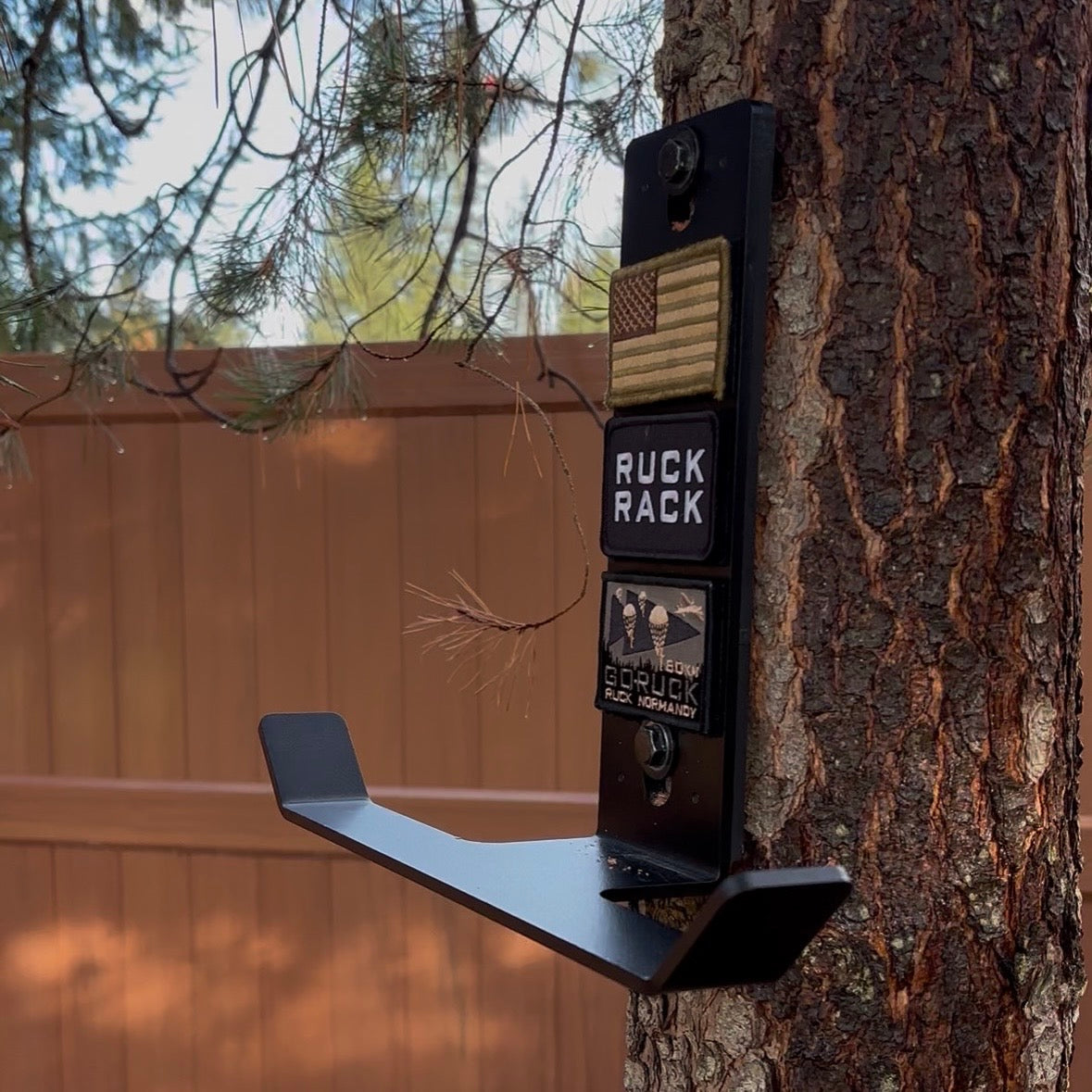 A black metal Ruck Rack Monster wall-mount hook attached to a tree trunk, displaying three tactical morale patches.