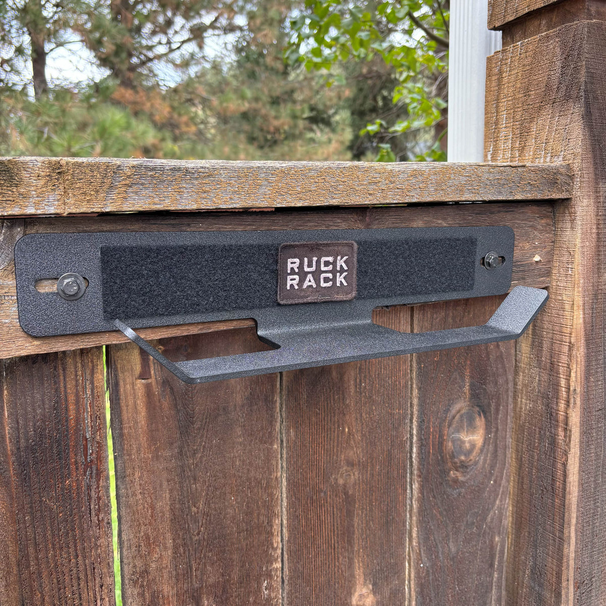 Black metal Ruck Rack wall-mounted storage rack attached to a wooden fence, featuring a central patch labeled “RUCK RACK.”