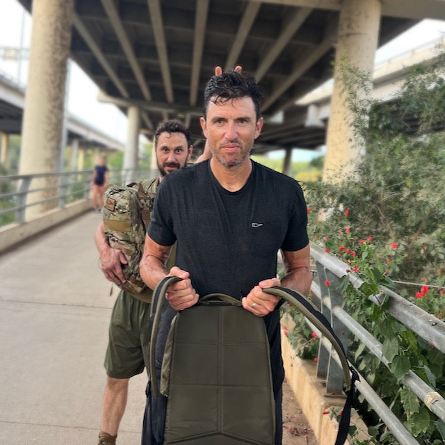 A man in a black t-shirt holds a backpack with classic simplicity, while a man in military-style gear stands behind him making bunny ears with his fingers.
