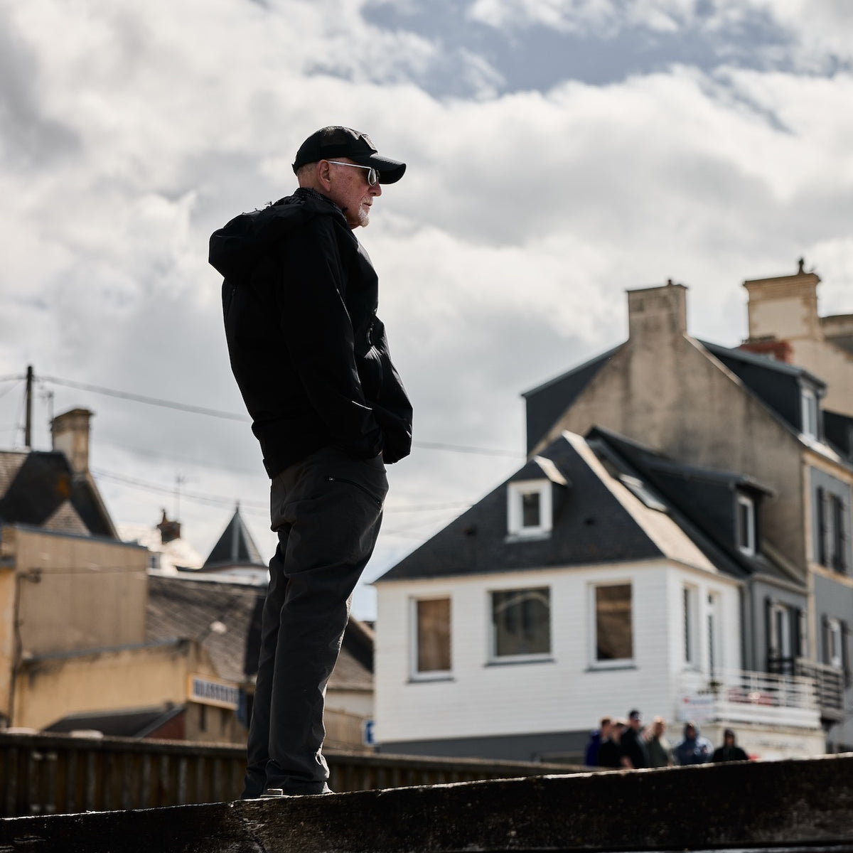 A man in a black hooded jacket and baseball cap stands on a ledge in tough technical pants, looking out over a town.
