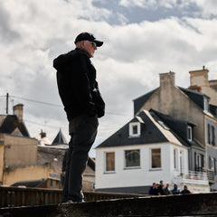 A man in a black hooded jacket and baseball cap stands on a ledge in tough technical pants, looking out over a town.