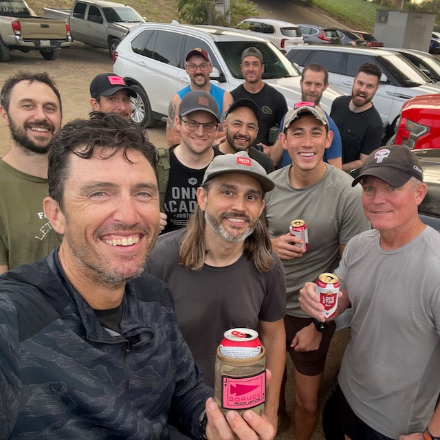 A large group of men smiling and holding cans of beer in Ballistic Beer Jackets while standing in an outdoor parking lot.