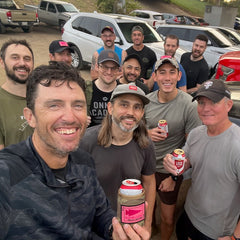 A large group of men smiling and holding cans of beer in Ballistic Beer Jackets while standing in an outdoor parking lot.
