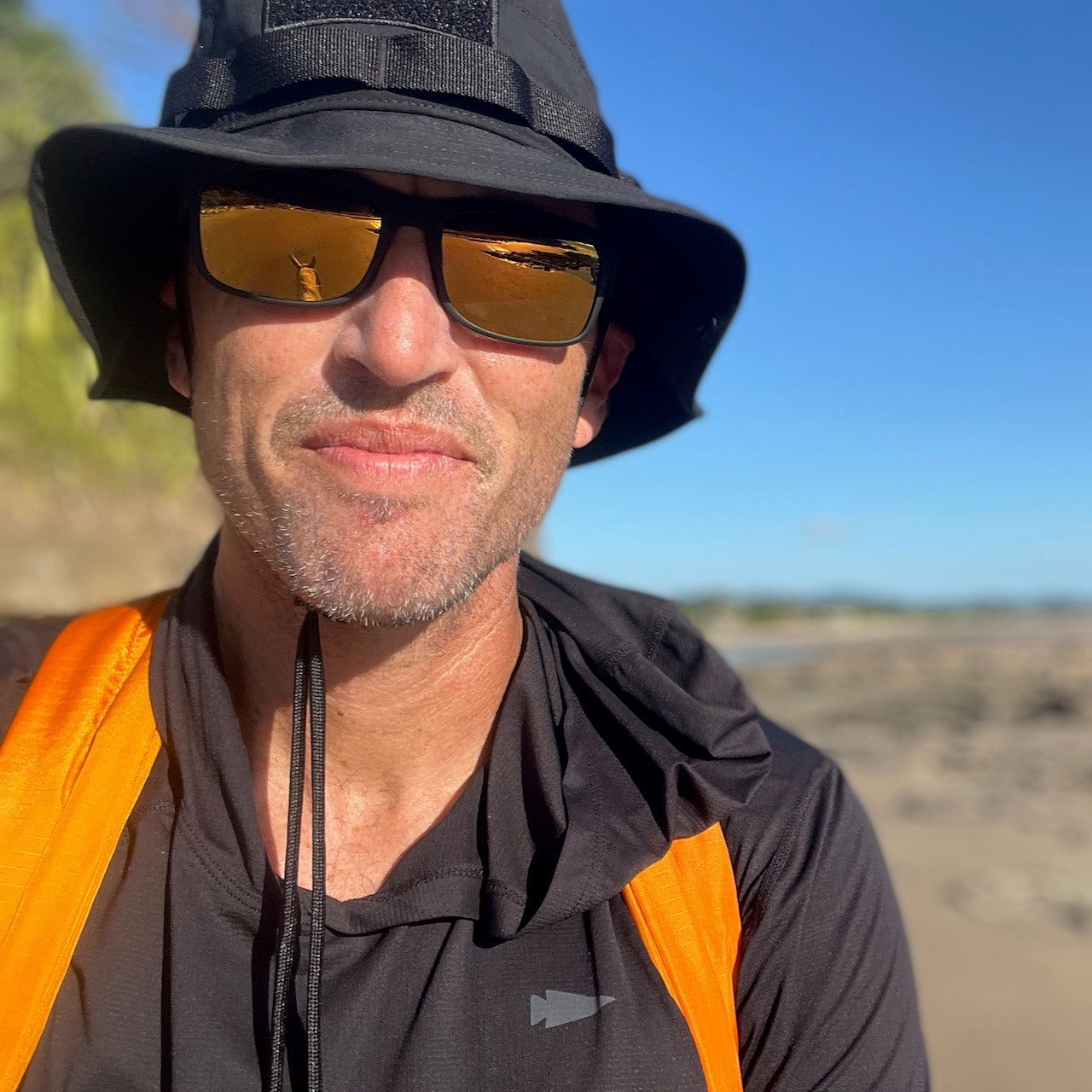 A man wearing a black hat, sunglasses, and the Packable Bullet Ruck - Ripstop ROBIC® in orange stands outdoors on a sunny day, surrounded by blue sky and rocky terrain.