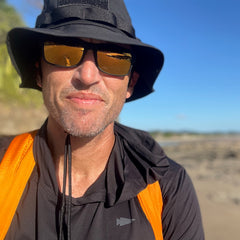 A man wearing a black hat, sunglasses, and the Packable Bullet Ruck - Ripstop ROBIC® in orange stands outdoors on a sunny day, surrounded by blue sky and rocky terrain.