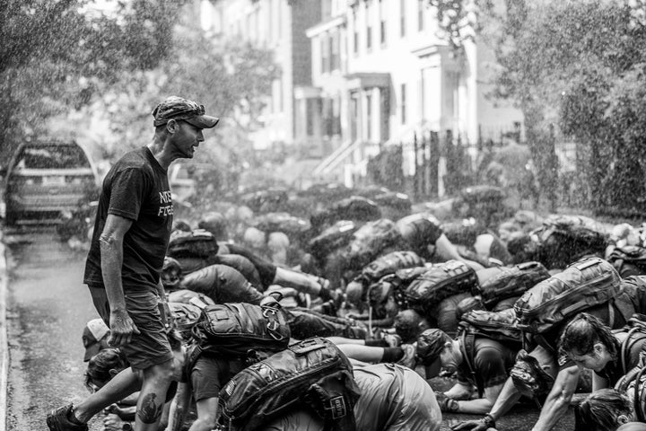 A group of people with backpacks crouch on a rainy street as a man stands nearby, instructing them.