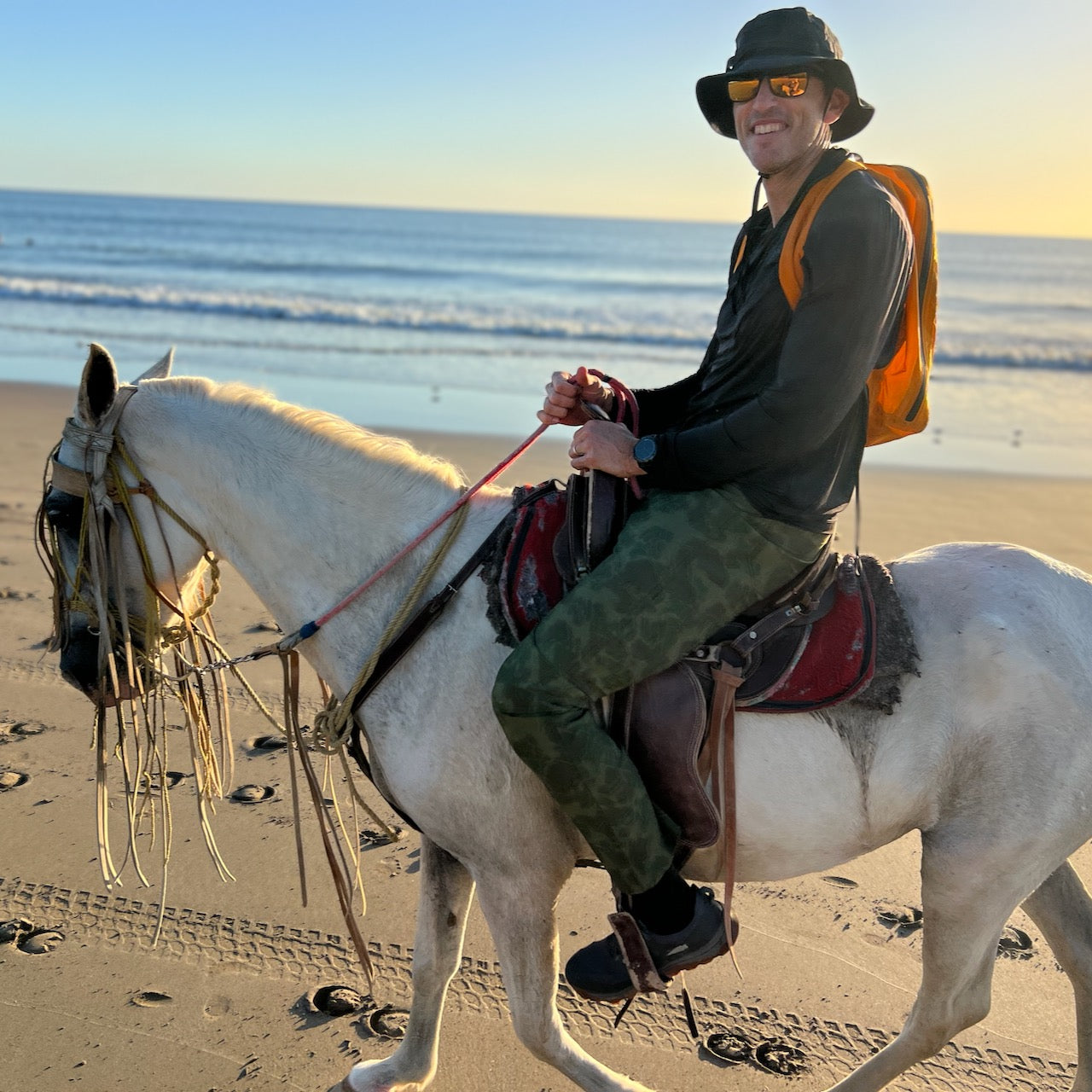 A person wearing the Boonie Hat - Tactical - ToughDry and sunglasses rides a white horse along a sandy beach by the ocean at sunset.