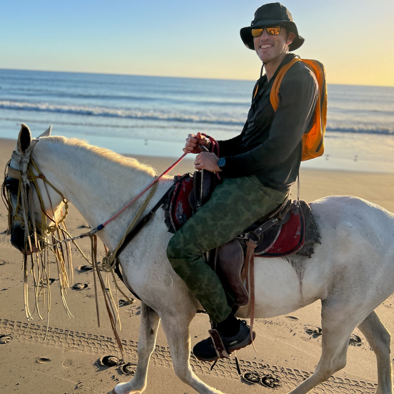 A smiling person in a hat and sunglasses rides a white horse along a sandy beach at sunset, carrying the Packable Bullet Ruck - Ripstop ROBIC® with AquaGuard zippers slung over their shoulder.