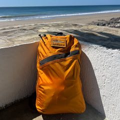 A Packable Bullet Ruck - Ripstop ROBIC® backpack rests against a white wall near a sandy beach and the blue ocean beneath a sunny sky.