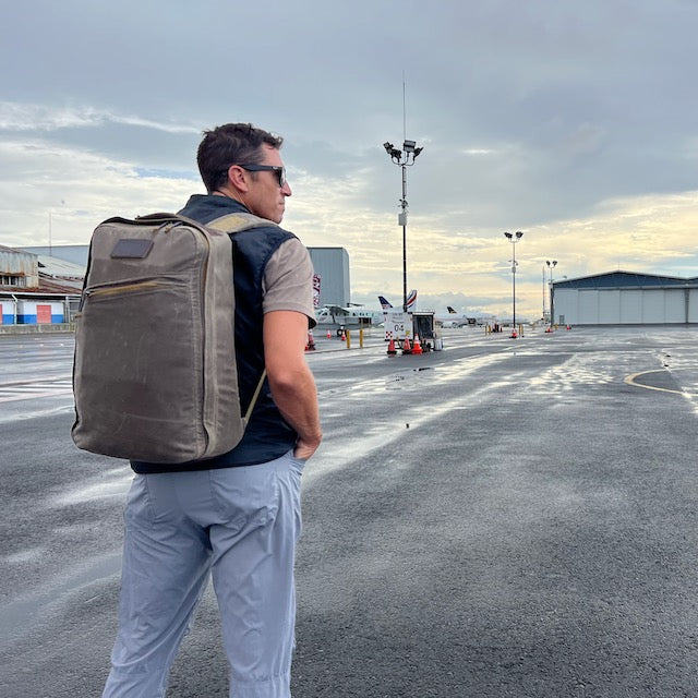 A man wearing sunglasses stands on a wet airport tarmac near hangars on a cloudy day, carrying the GR1 USA - Heritage Waxed Canvas backpack.