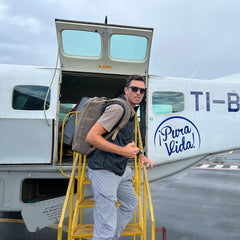 Man with sunglasses and a GR1 USA - Heritage Waxed Canvas backpack boards a small airplane with "Pura Vida!" written on the side.