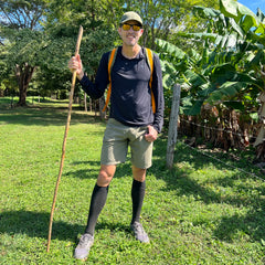 Man in outdoor gear stands on grass with a walking stick, wearing sunglasses, a backpack, and the Performance Running Hat - Italian ToughMesh made from premium Italian fabric.