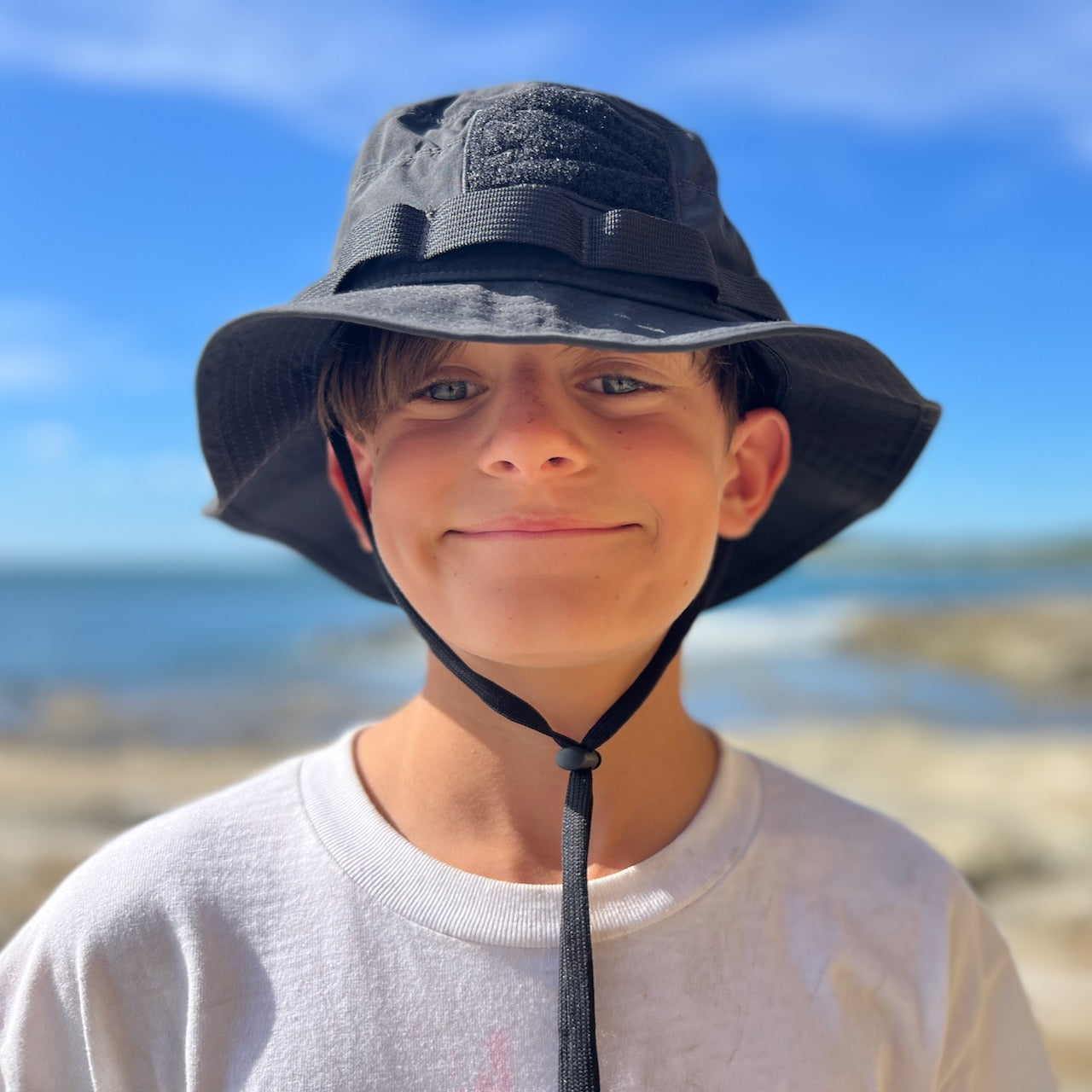 Smiling boy in a white shirt wearing the Boonie Hat - Tactical - ToughDry, standing outdoors by the beach on a sunny day.