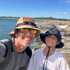 A man and a boy wearing Boonie Hat - Tactical - ToughDry smile by the rocky seaside under a clear blue sky.