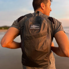 A man stands outdoors in profile with a blurred beach background, wearing the Packable Bullet Ruck - Ripstop ROBIC® in gray.