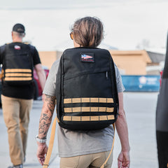 A person with a tattooed arm is wearing a black GR1 rucksack while walking outdoors with others dressed similarly.