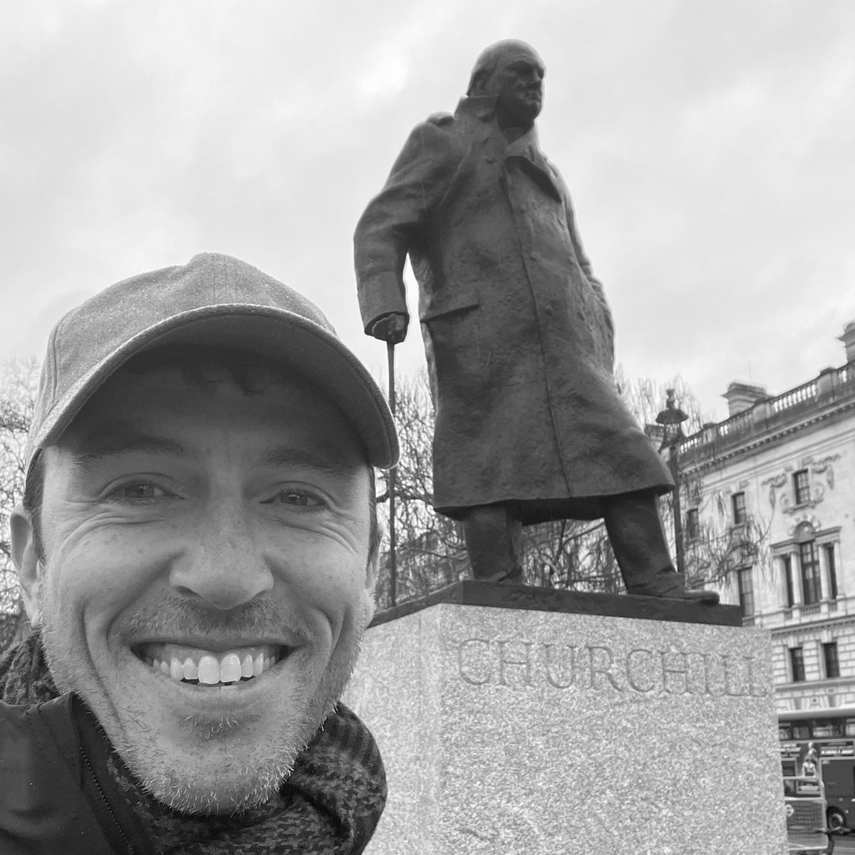 A smiling person wearing the Tac Hat - Heritage poses in front of a large statue labeled “CHURCHILL” in a city square.