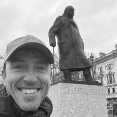 A smiling person wearing the Tac Hat - Heritage poses in front of a large statue labeled “CHURCHILL” in a city square.