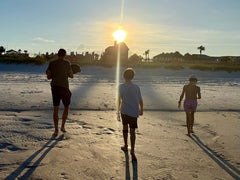 Three people walk on a sandy beach at sunset, casting long shadows toward the shoreline and houses—a peaceful moment captured in the spirit of remembrance, much like the Monster Forever Tee - PREORDER honors beloved pets waiting beyond the Rainbow Bridge.