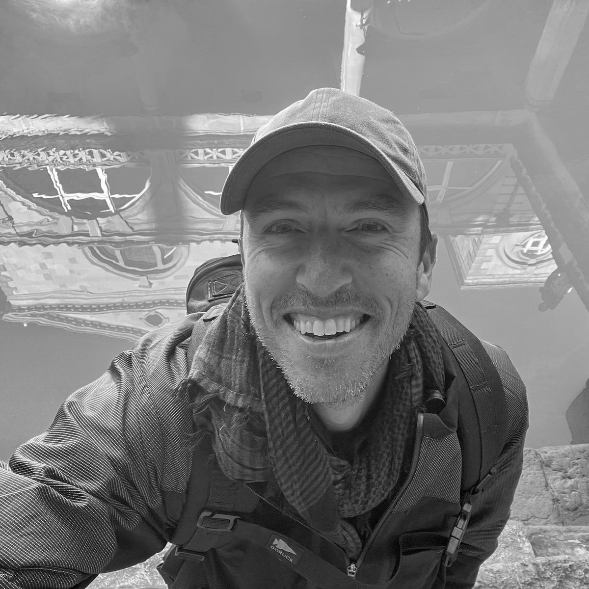 A man wearing the Tac Hat - Heritage and a jacket smiles for a selfie with reflections of buildings in the water behind him.
