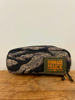 The Shadow Pocket- Tiger Stripe is a camo-patterned pouch made from rugged 1000D Cordura, featuring an "EMBRACE THE SUCK" patch. Ideal for rucksack organization, it’s displayed on a wooden surface.