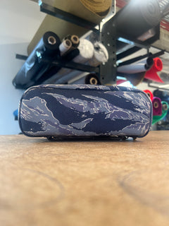 The Shadow Pocket- Tiger Stripe, a blue Cordura bag ideal for rucksack organization, rests on a wooden table with softly blurred fabric rolls in the background.