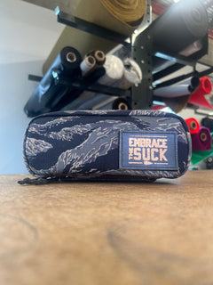 The Shadow Pocket- Tiger Stripe is a fabric pouch featuring a "EMBRACE THE SUCK" patch—ideal for organizing your rucksack—with rolls of fabric in the background.