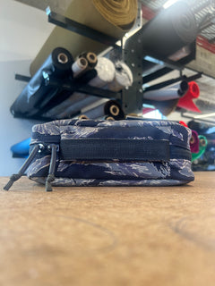 A Shadow Pocket- Tiger Stripe zippered pouch sits on a table, ideal for ruck organization, with rolls of fabric neatly stacked on shelves in the background.