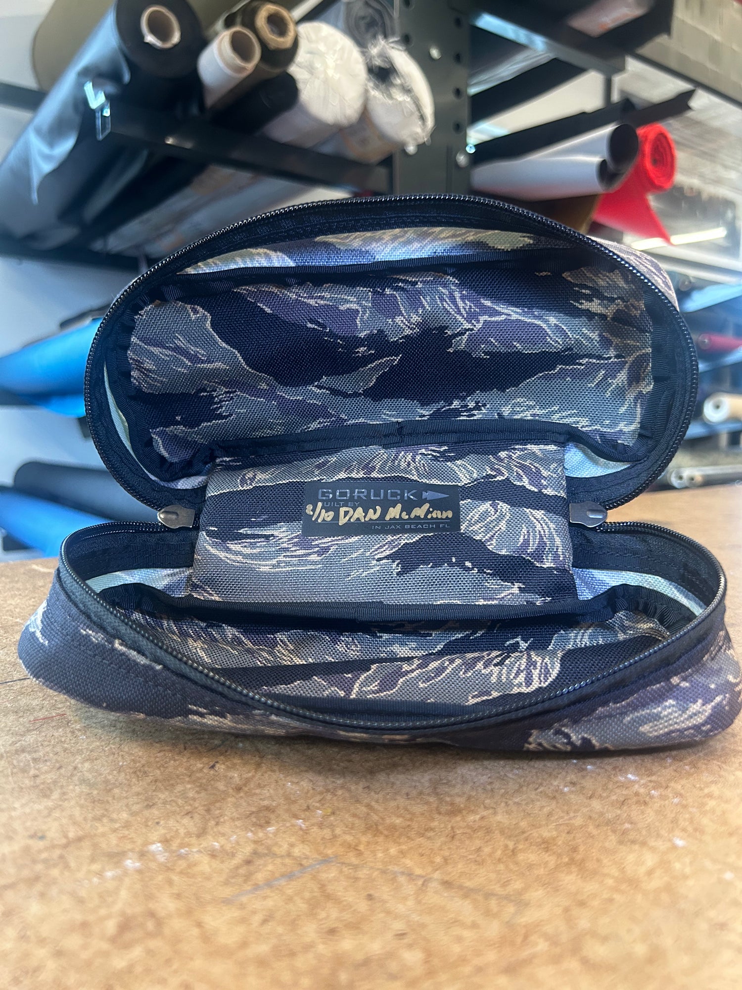 A Tiger Stripe Shadow Pocket pencil case sits open on a wooden surface, ideal for ruck organization, with shelves filled with rolls visible in the background.