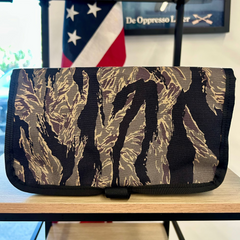 Scars Wire Dopp travel organizer in camouflage print sits on a wooden shelf with a blurred U.S. flag backdrop—ideal for keeping your EDC essentials safe and organized.
