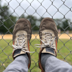 A person wearing Mackall 45 Fog sneakers and gray pants rests their feet on a chain-link fence by a baseball field.