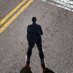 Shadow of a person standing on asphalt near yellow road lines, with their legs and Mackall 45 Fog shoes visible at the bottom.