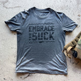 The Embrace the Suck Tee - Tri-Blend, featuring the iconic USMC motto in bold print, is displayed flat on a concrete surface.