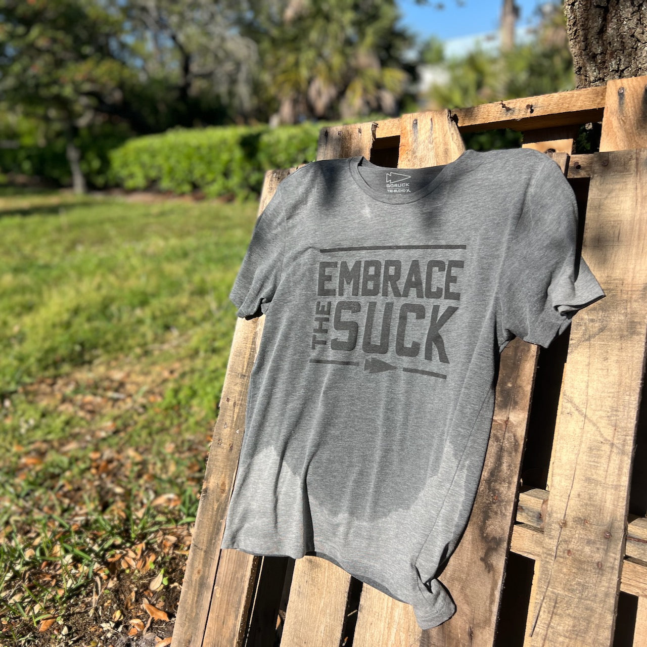 Embrace the Suck Tee - Tri-BlendX, featuring the iconic USMC motto, displayed outdoors on a wooden pallet in sunlight.