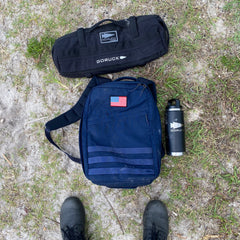 A pair of boots, a black GORUCK bag, a US flag-patched navy backpack, and Simple Training Sandbags with a GORUCK bottle sit on the grass—ideal for sandbag training.