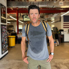 Wearing the Three Rules Tee - Unisex Tri-BlendX, a man with a backpack and wet t-shirt stands indoors in front of an American flag, showing you can look cool and confident anywhere.