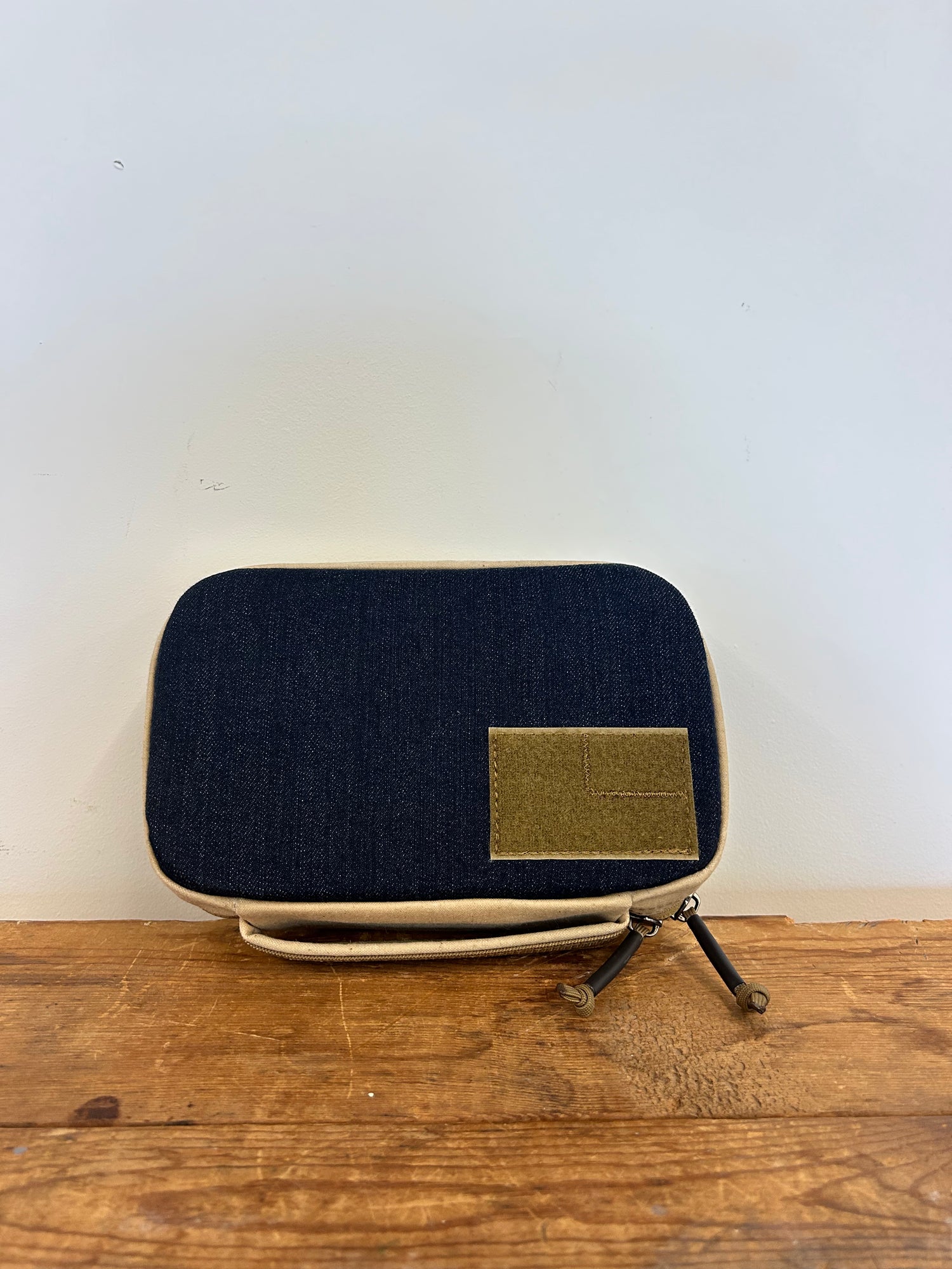 The GR1 Field Pocket - Denim and Waxed Canvas, a rectangular zippered pouch in navy blue and beige with a brown patch, is displayed on a wooden surface.