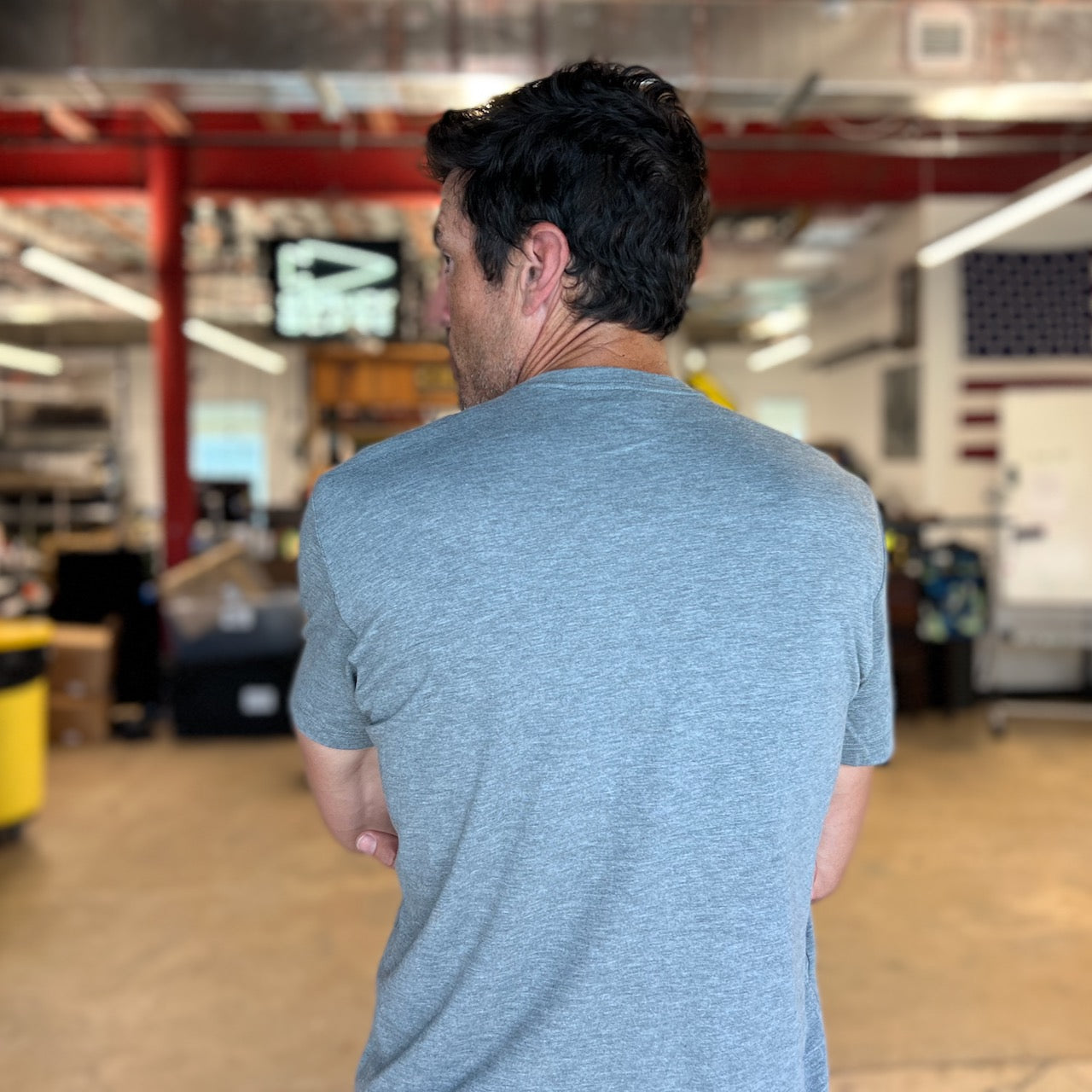 A man is seen indoors with his back to the camera, wearing the Subtle Spearhead Tee - Unisex Tri-Blend in gray, inside a spacious room with an industrial vibe.