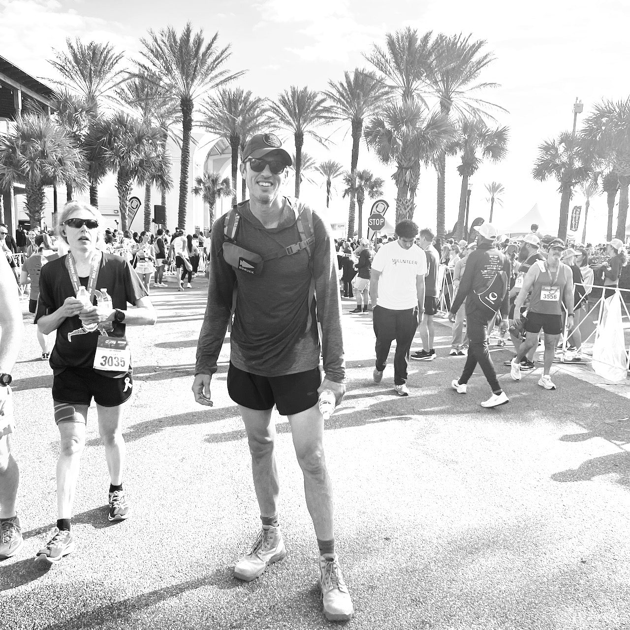 Runners celebrate at a palm-lined finish area; in front, a smiling man wears race gear, a medal, and the Sternum Pouch - Cordura.