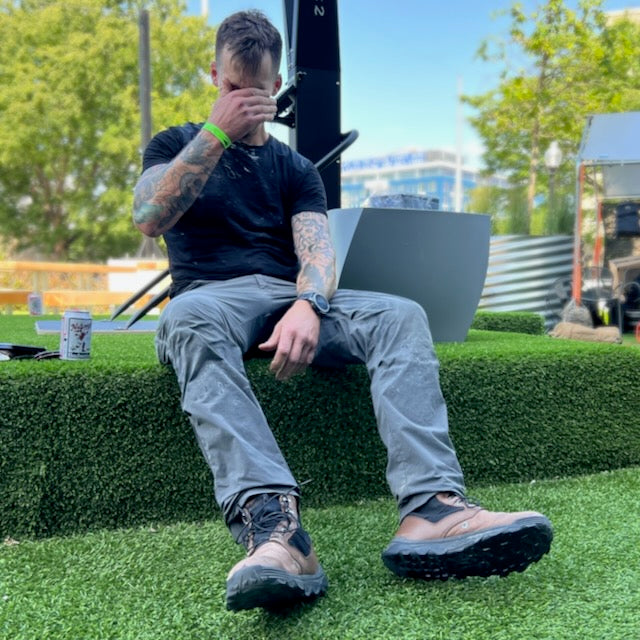 A tattooed man sits on artificial grass outdoors, covering his face with one hand, wearing the Men’s Commando Tee made from Merino Wool.