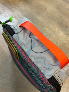Close-up of the Ripstop Robic XL Packing Cube in gray, made from durable nylon with a bright orange handle and green zipper pull—perfect for travel organization or use with other packing cubes, shown on a textured floor.