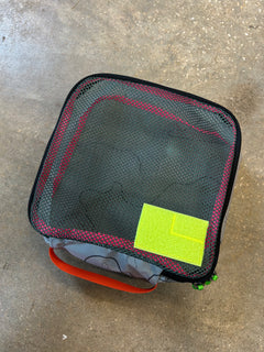 The Ripstop Robic XL Packing Cube, featuring a black mesh top, red trim, orange handle, and bright green label, rests on concrete—perfect for keeping your travel essentials organized.