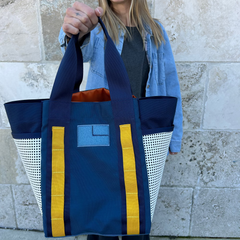 Smiling woman holding the Scars Tote toward the camera in front of a stone wall.